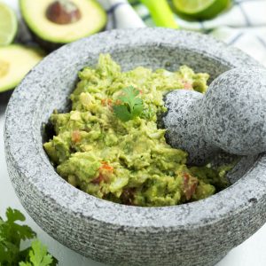 guacamole in a mortary with a spring of cilantro on top. Avocado, cilantro and lemon on the table next to the bowl.