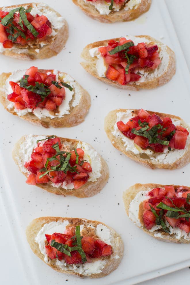 multiple slices of fresh bread with goat cheese and chipped strawberries and basil sitting on a white rectangular plate