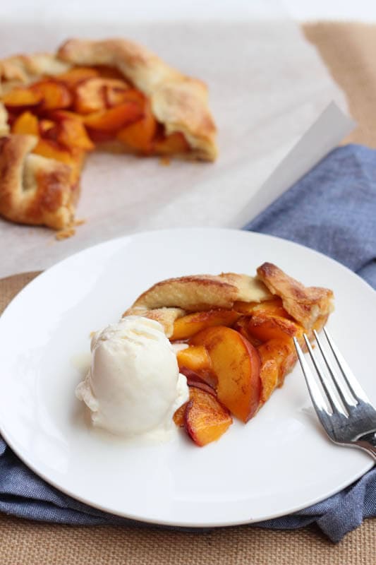 A slice of peach tart sitting on a white plate with one scoop of vanilla ice cream and a fork.