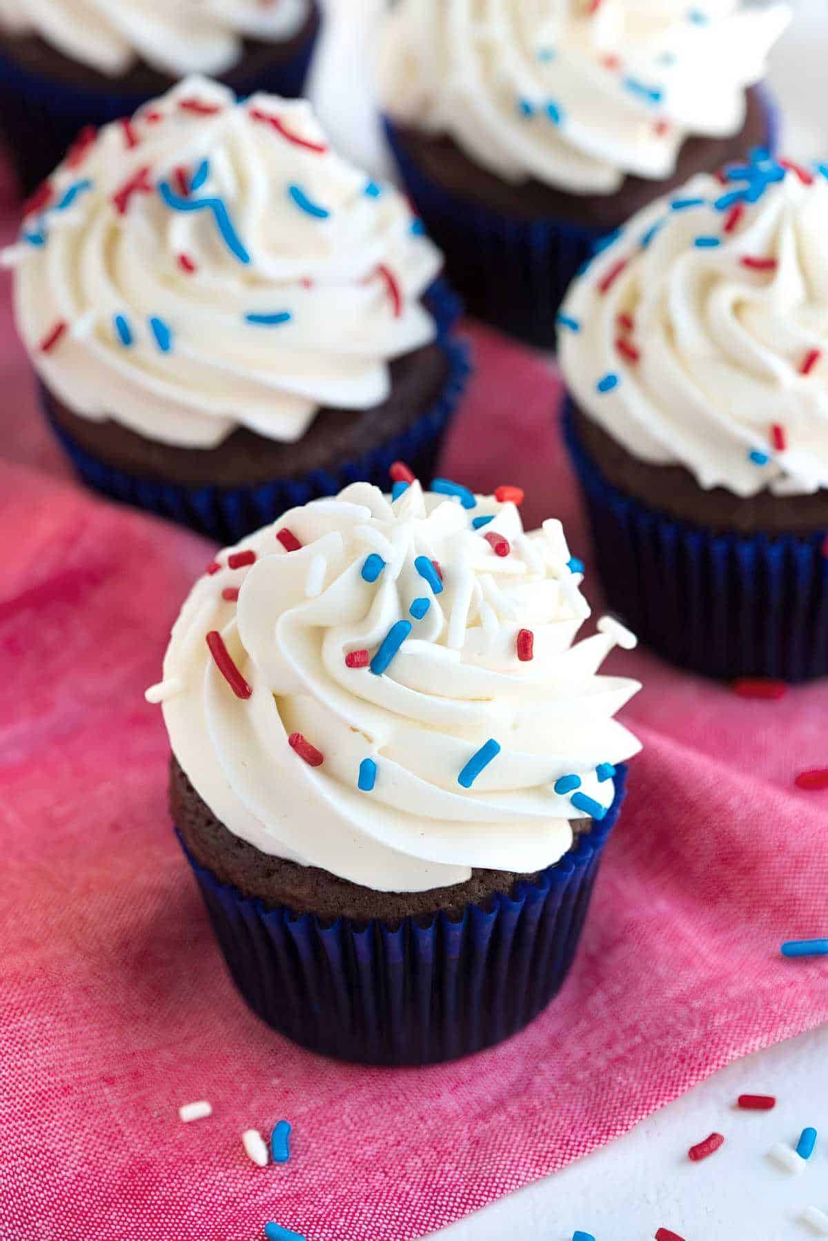 Chocolate cupcakes topped with vanilla buttercream frosting and red, white, and blue sprinkles on a pink cloth.