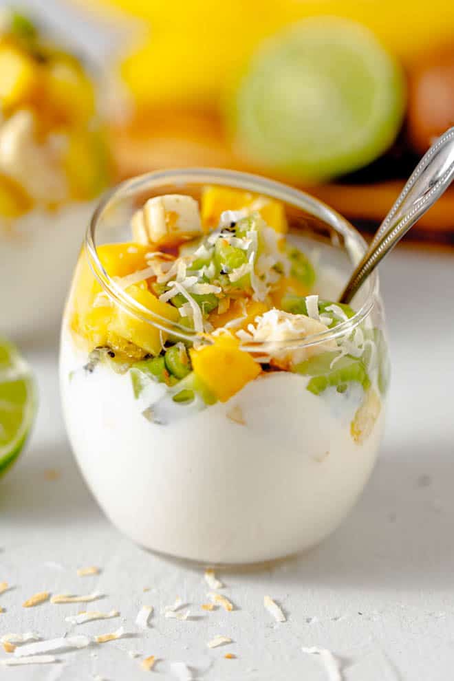 clear glass bowl with greek yogurt and mango, banana and kiwi on top with toasted coconut.