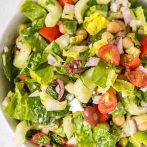 A white bowl filled with chopped Mediterranean salad including chicken peas, lettuce, tomatoes, olives and cucumbers.