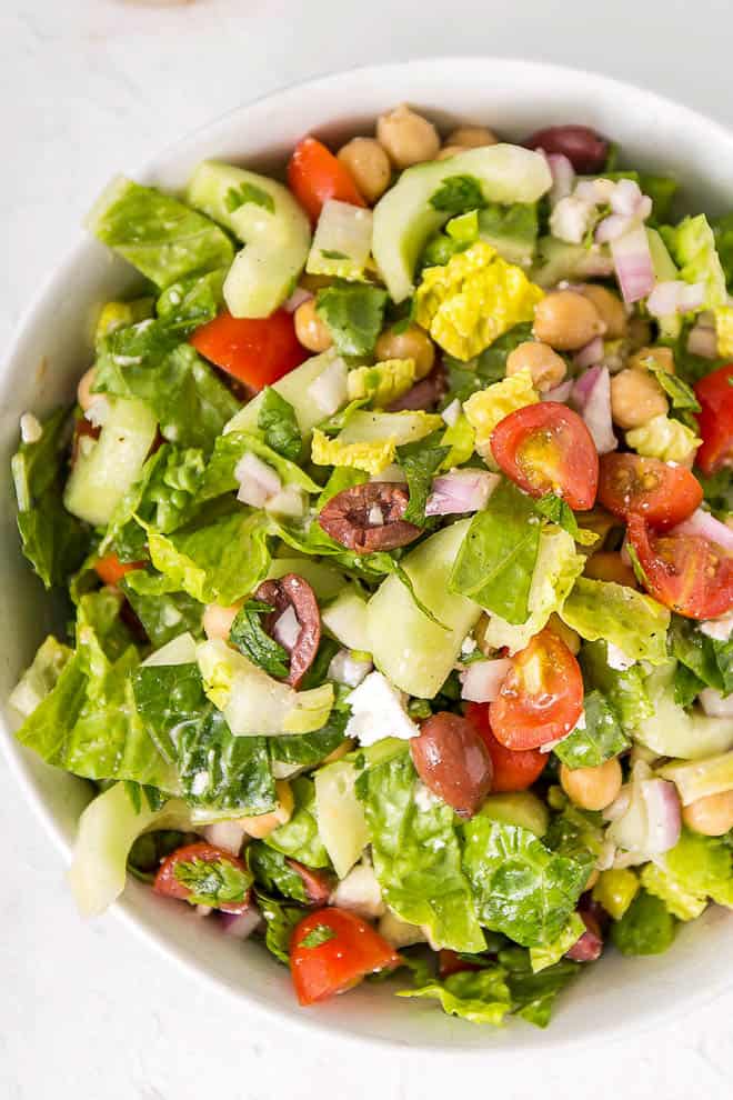 A white bowl filled with chopped Mediterranean salad including chicken peas, lettuce, tomatoes, olives and cucumbers.