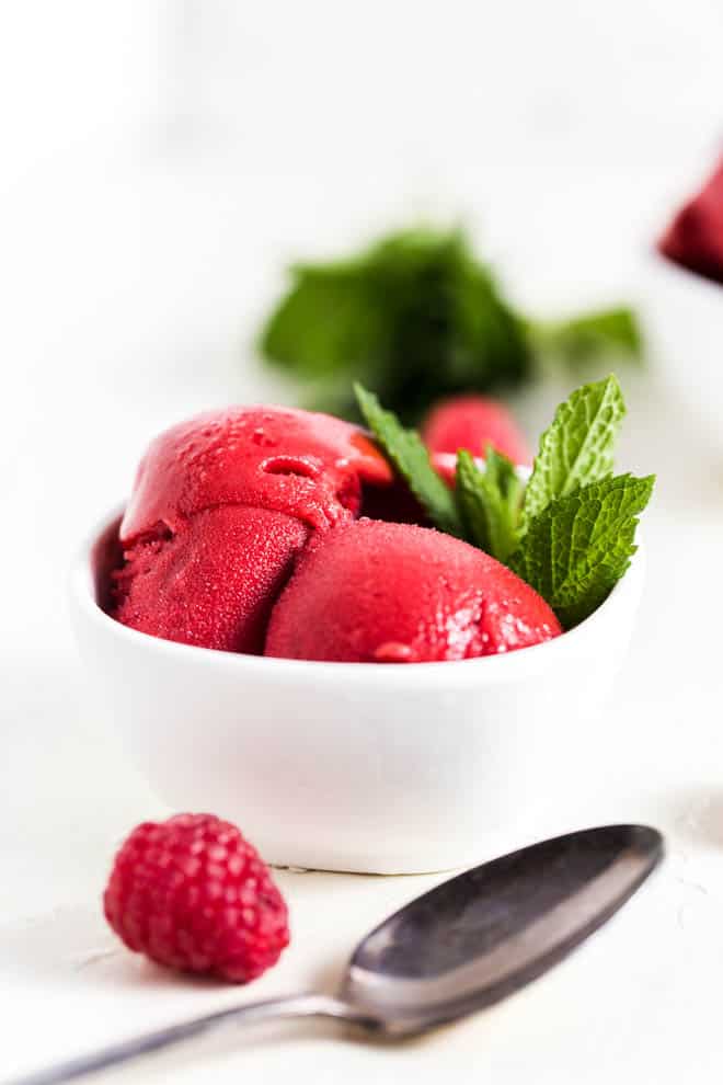 Two scoops of raspberry sorbet in a white bowl with mint leaves on the side and a raspberry sitting in front of the bowl.