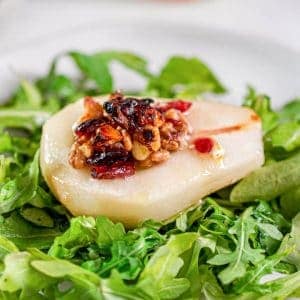 Roasted pear with blue cheese, cranberries and walnuts sitting on a bed of arugula on a plate.