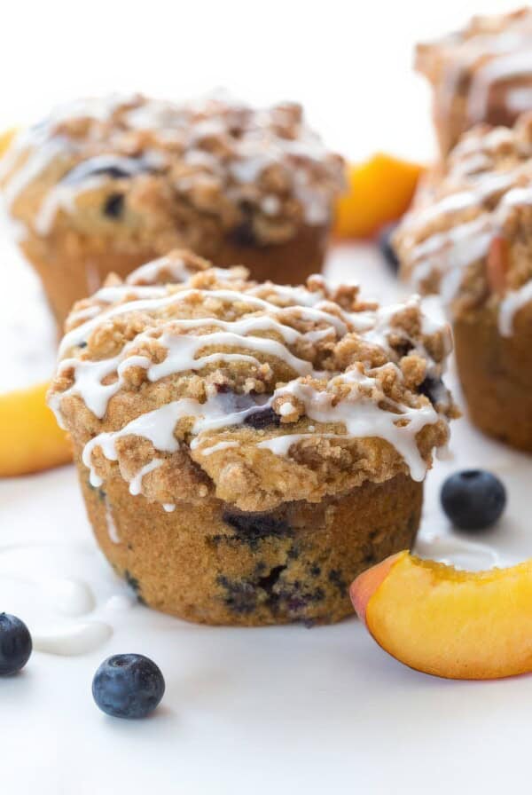 Close-up of a blueberry peach cobbler muffin topped with streusel and icing drizzle, surrounded by fresh blueberries and peach slices.