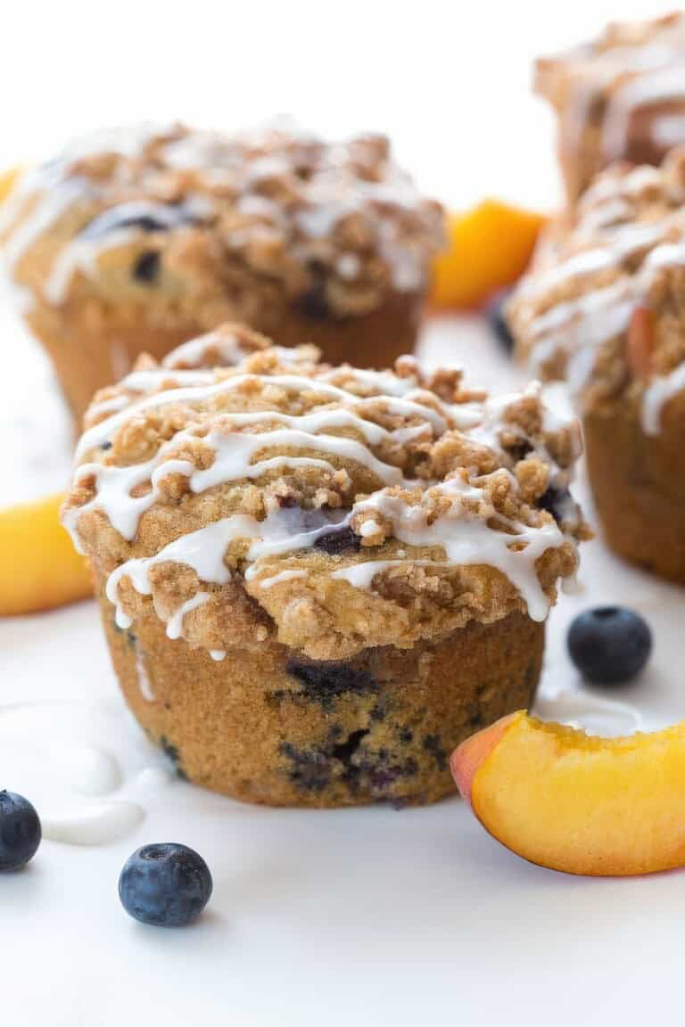Close-up of a blueberry peach cobbler muffin topped with streusel and icing drizzle, surrounded by fresh blueberries and peach slices.