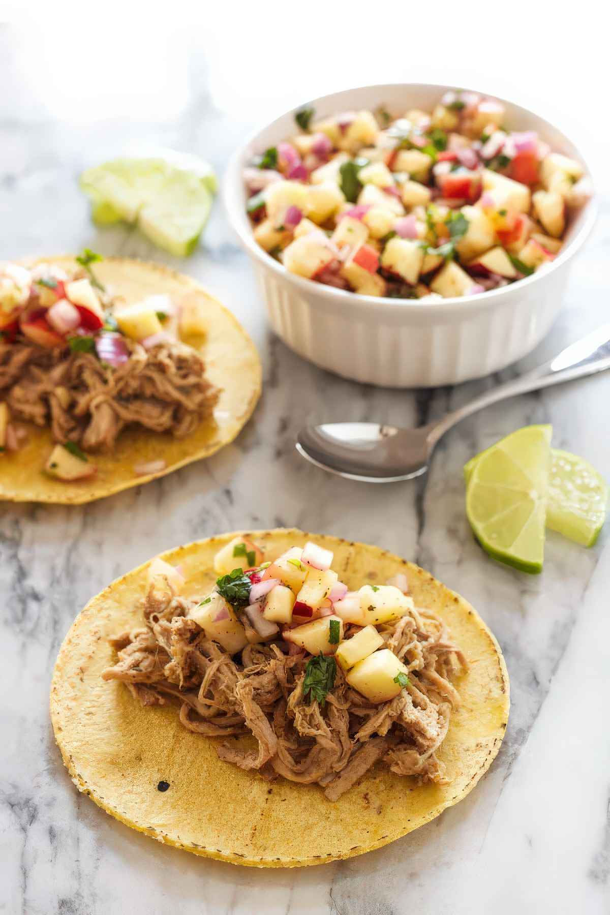 Slow cooker carnitas tacos topped with fresh apple salsa, with extra salsa and lime wedges on the side.