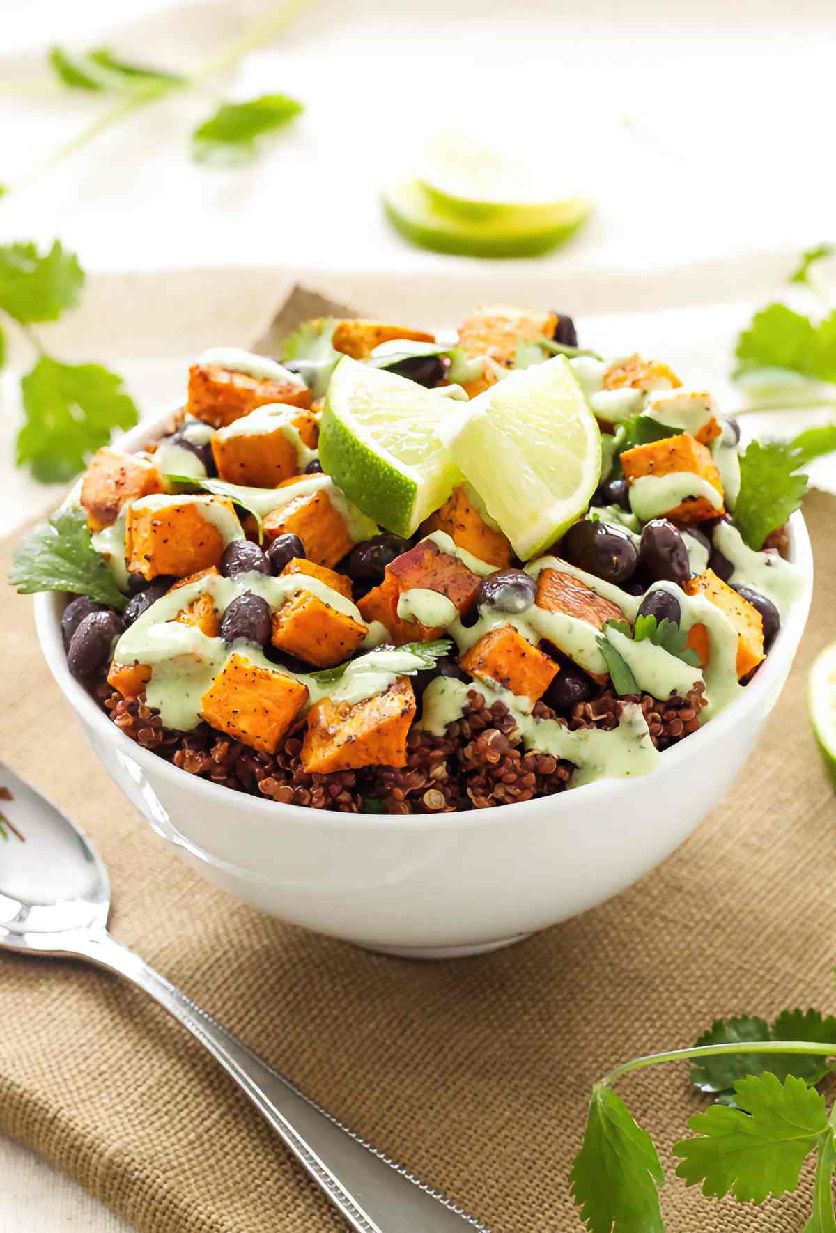 White bowl filled with red quinoa, roasted sweet potatoes and black beans, finished with cilantro cream and lime wedges.