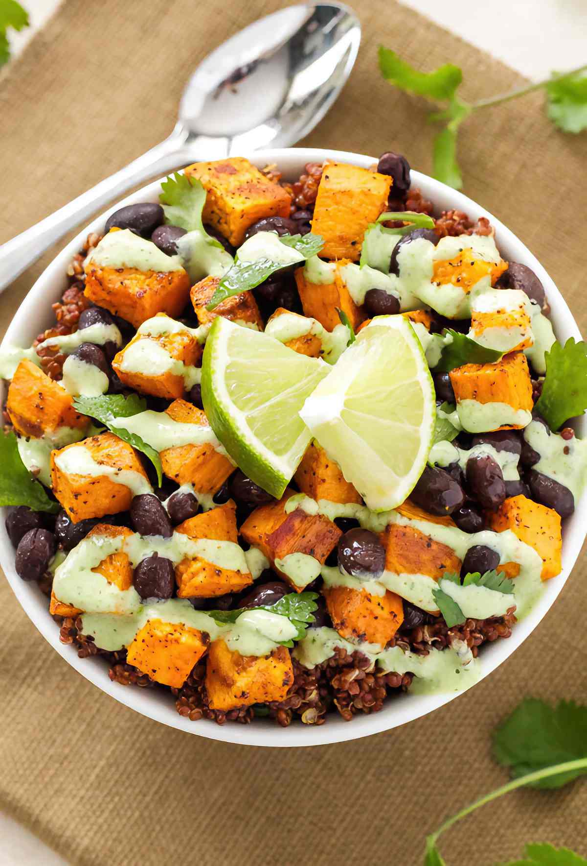 Overhead view of a sweet potato and black bean quinoa bowl with cilantro leaves, green cilantro cream and lime wedges.