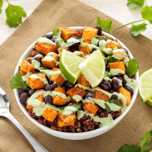 Sweet potato and black bean quinoa bowl topped with cilantro cream and lime wedges, with a spoon alongside.