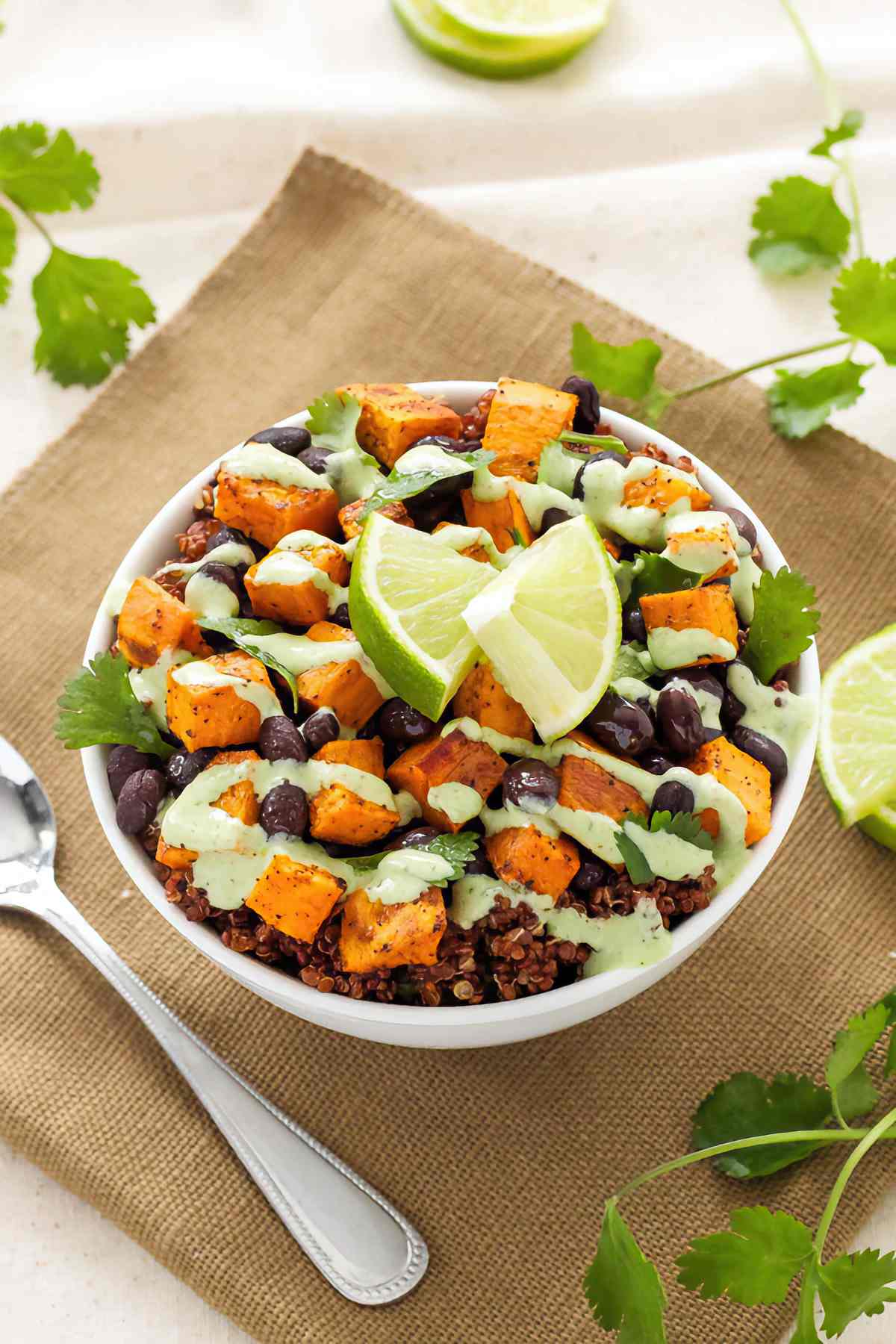 Sweet potato and black bean quinoa bowl topped with cilantro cream and lime wedges, with a spoon alongside.