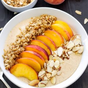 Peach pie smoothie in a white bowl topped with freshly sliced peaches, granola and sliced almonds.
