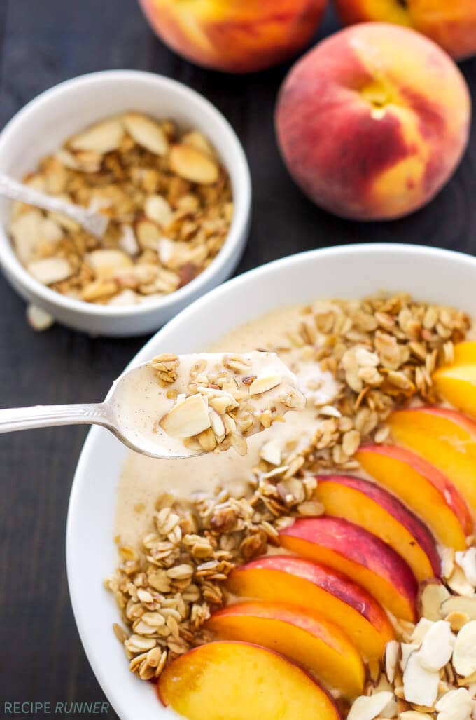 A spoon scooping into a bowl of peach smoothie with toppings like fresh sliced peaches and sliced almonds.