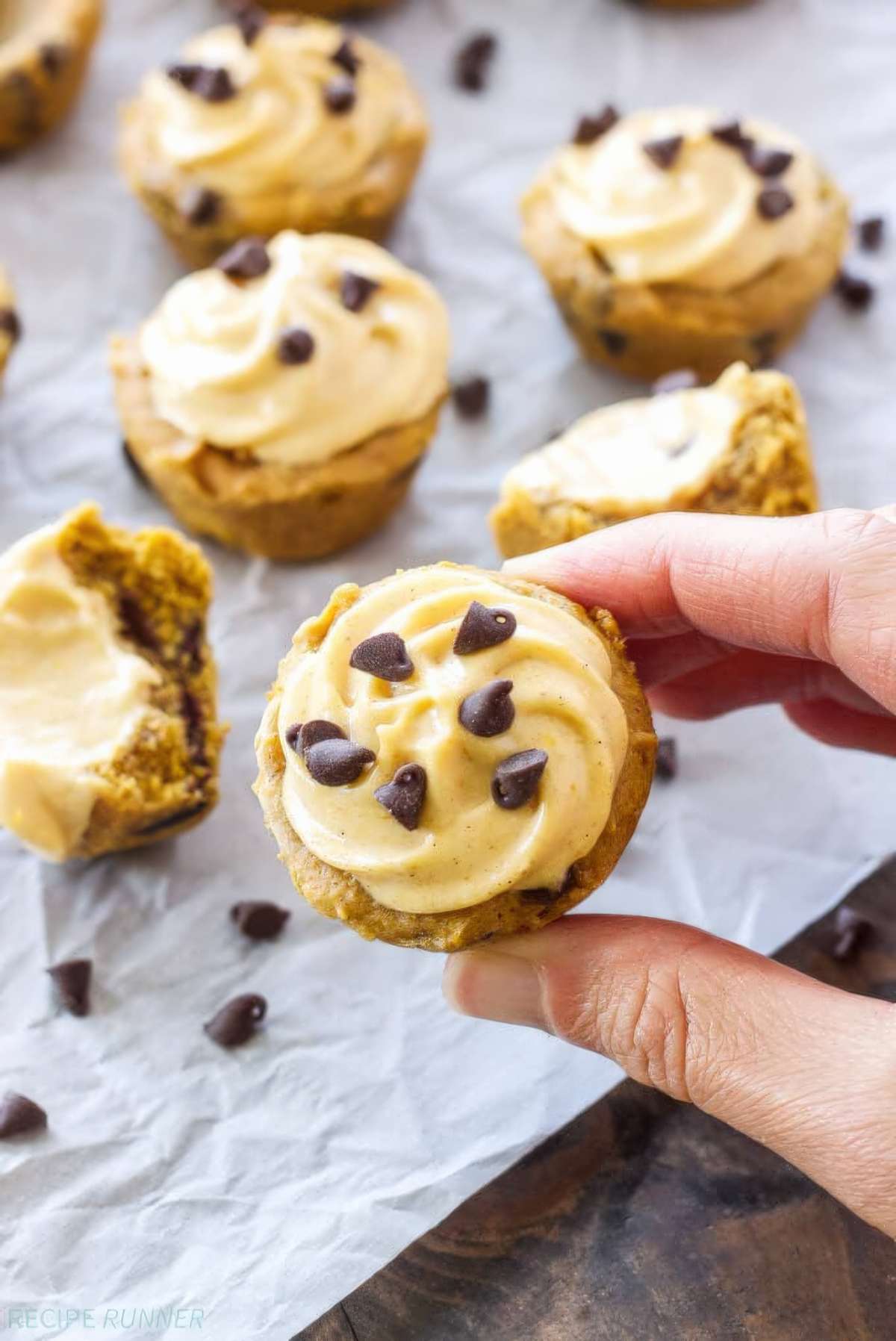 A hand holds a pumpkin chocolate chip cookie cup topped with pumpkin cream cheese frosting, showing its small size.