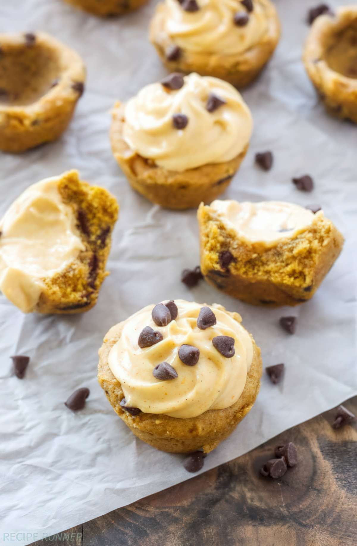 Halved pumpkin chocolate chip cookie cups reveal the soft cookie centers and pumpkin cream cheese frosting alongside whole frosted cups.
