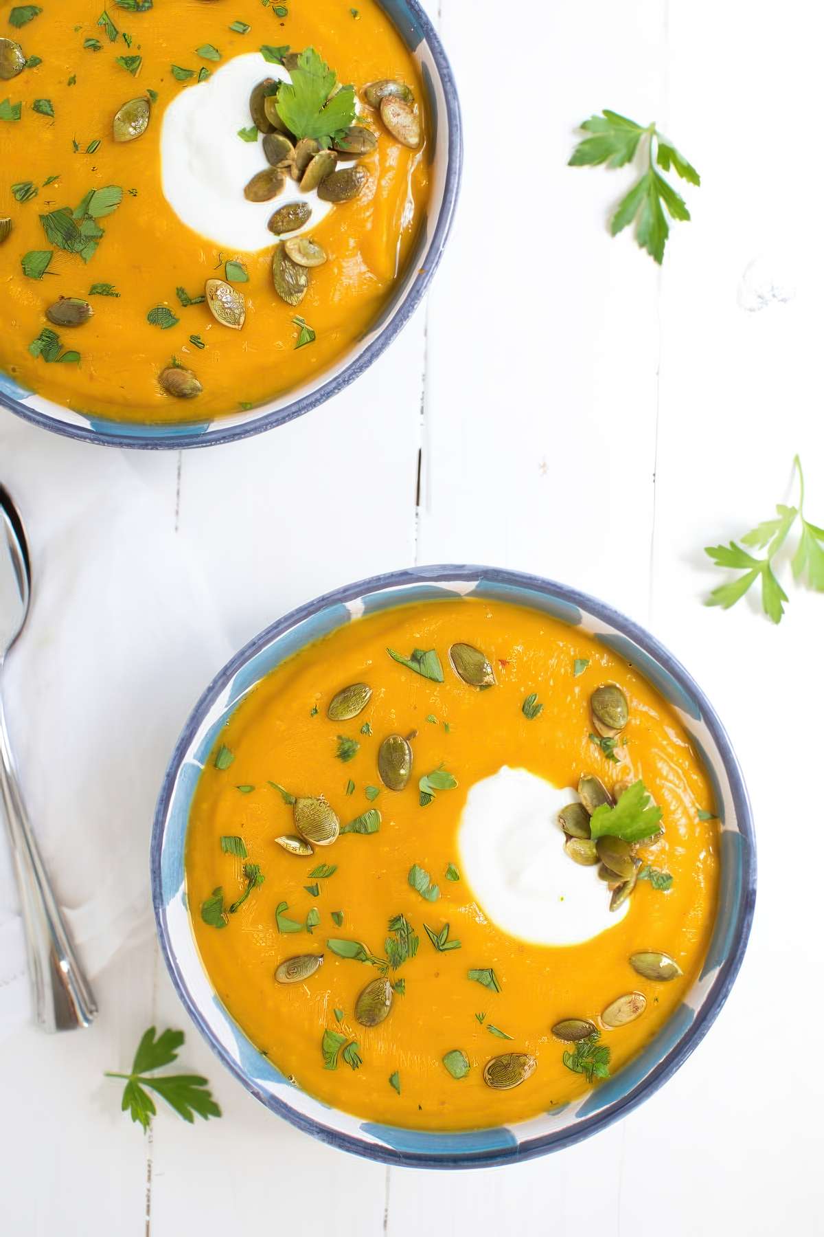 Two bowls of roasted butternut squash and red lentil soup topped with yogurt, pepitas and parsley on a white wooden table.