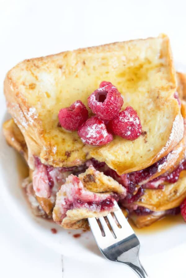 Close-up of a forkful of raspberry cheesecake-stuffed brioche French toast, showing the creamy filling and raspberry preserves, topped with fresh raspberries and powdered sugar.