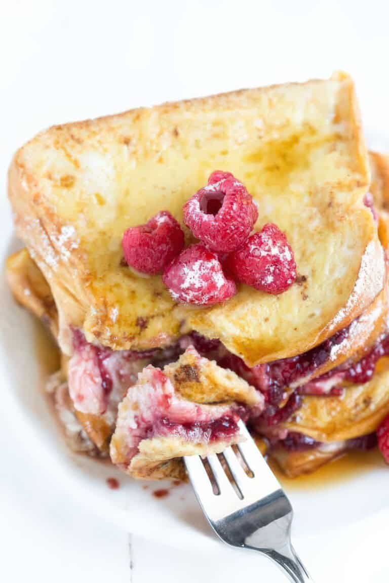 Close-up of a forkful of raspberry cheesecake-stuffed brioche French toast, showing the creamy filling and raspberry preserves, topped with fresh raspberries and powdered sugar.