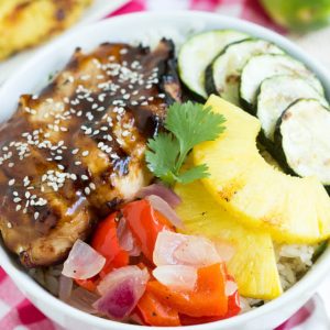 teriyaki chicken, grilled zucchini, pineapple and vegetables in a bowl over rice sitting on a pink checker napkin