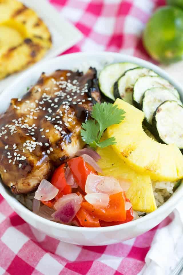 teriyaki chicken, grilled zucchini, pineapple and vegetables in a bowl over rice sitting on a pink checker napkin
