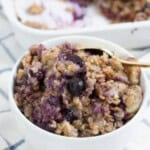 Close-up of a bowl filled with baked oatmeal, featuring blueberries and oats, with a gold spoon and baking dish in the background.