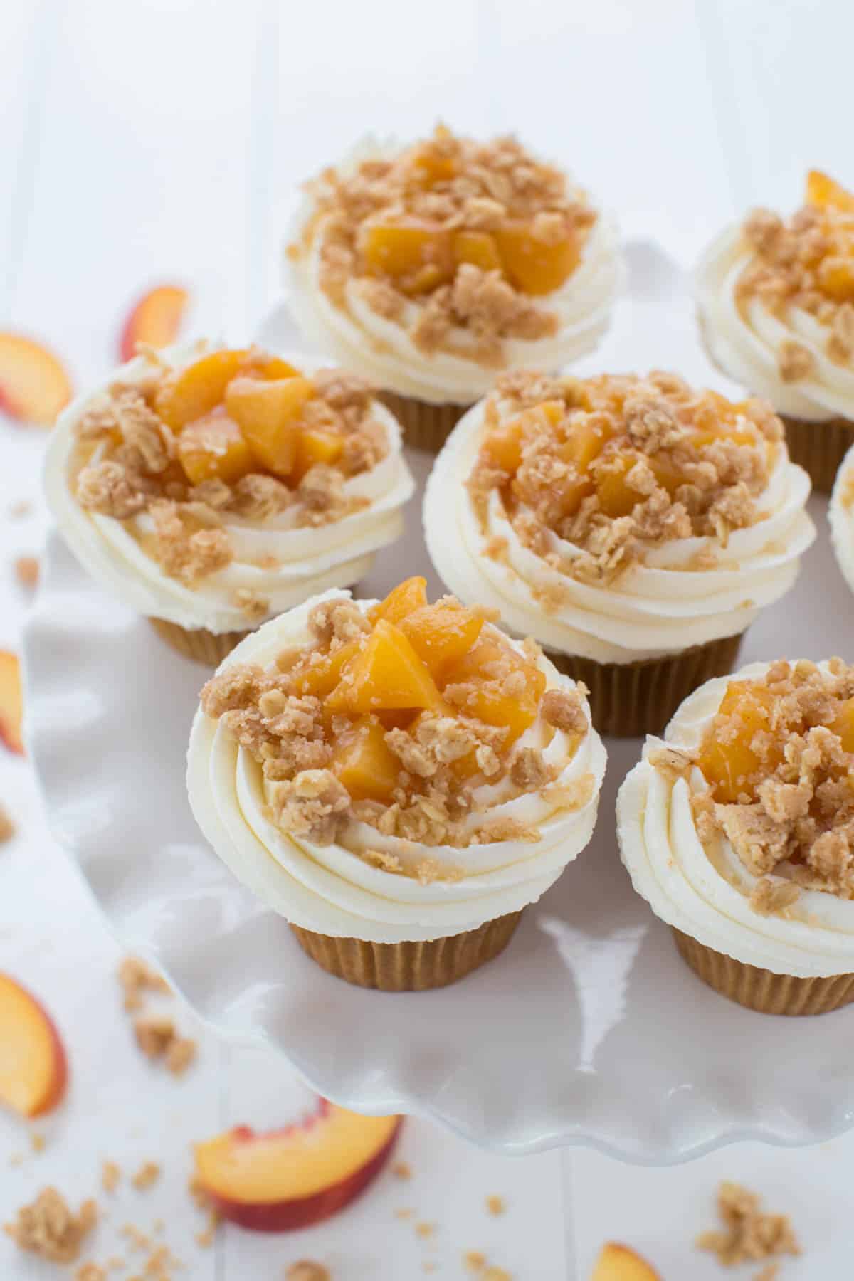 Peach pie cupcakes sit on top of a cupcake stand with slices of fresh peaches off to the side.