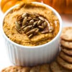 a white bowl with pumpkin hummus sitting next to a stack of crackers and two mini pumpkins