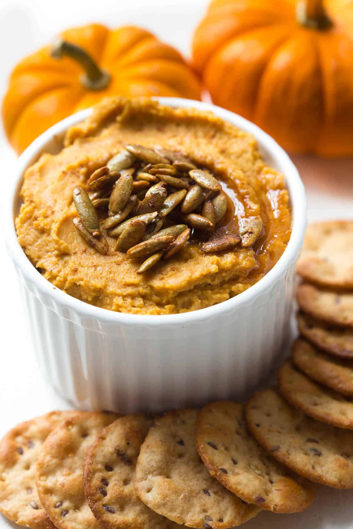 a white bowl with pumpkin hummus sitting next to a stack of crackers and two mini pumpkins