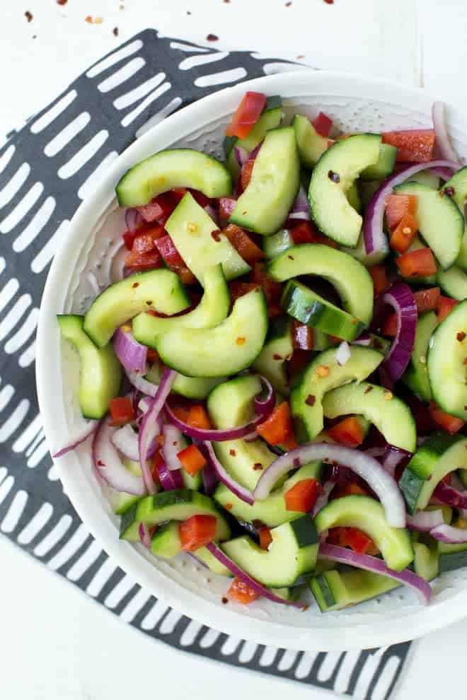 sliced cucumbers, red peppers and red onions in a white bowl sitting on a white background with a blue napkin