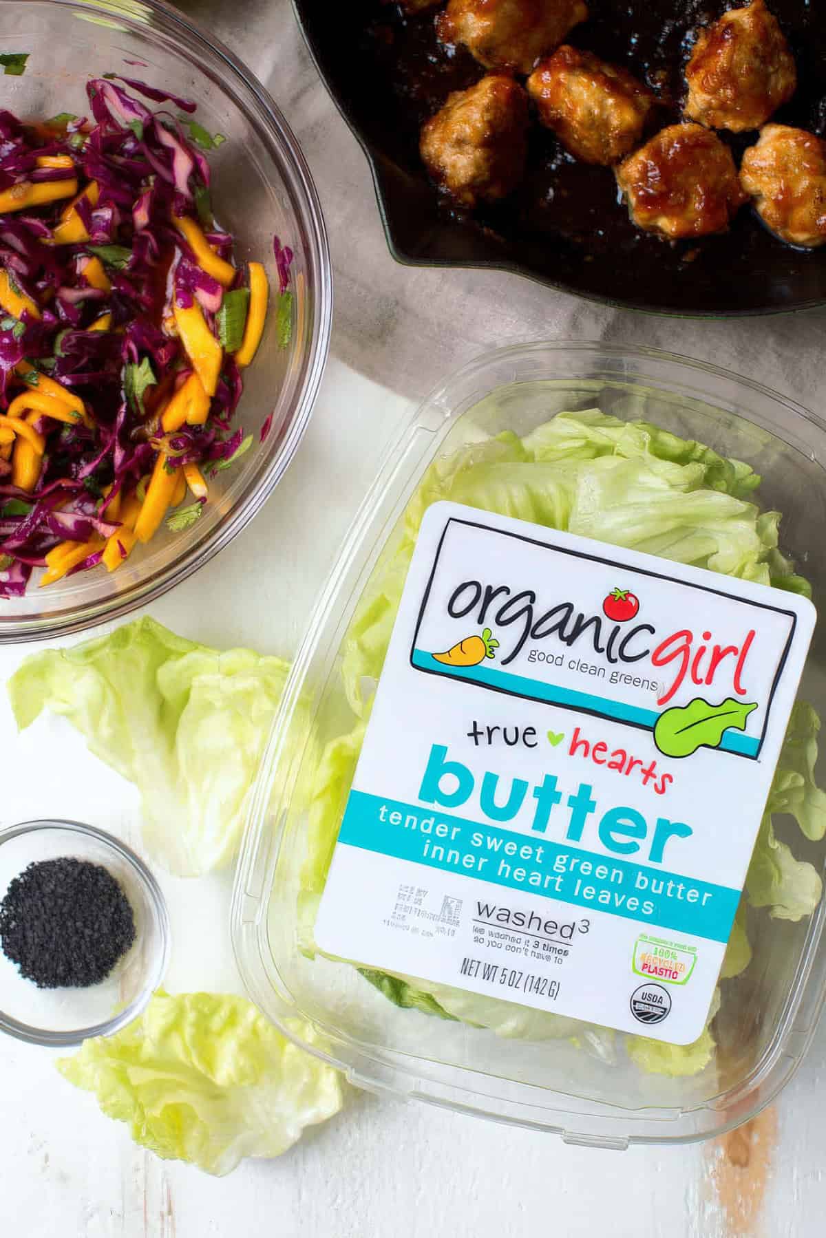 Overhead view of ingredients for lettuce wraps, including a container of butter lettuce, cooked glazed meatballs in a skillet, shredded red cabbage and sliced mango in a glass bowl, and a small bowl of black sesame seeds.