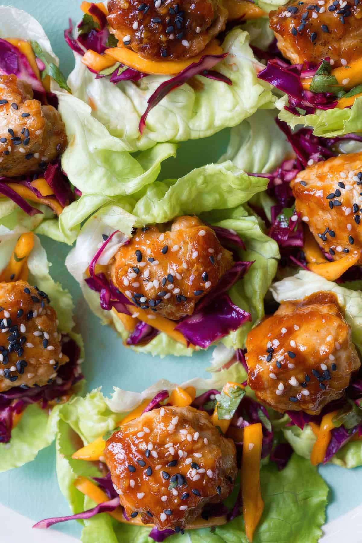 Overhead shot of glazed chicken meatballs on butter lettuce leaves, topped with shredded red cabbage, sliced mango, and black and white sesame seeds.