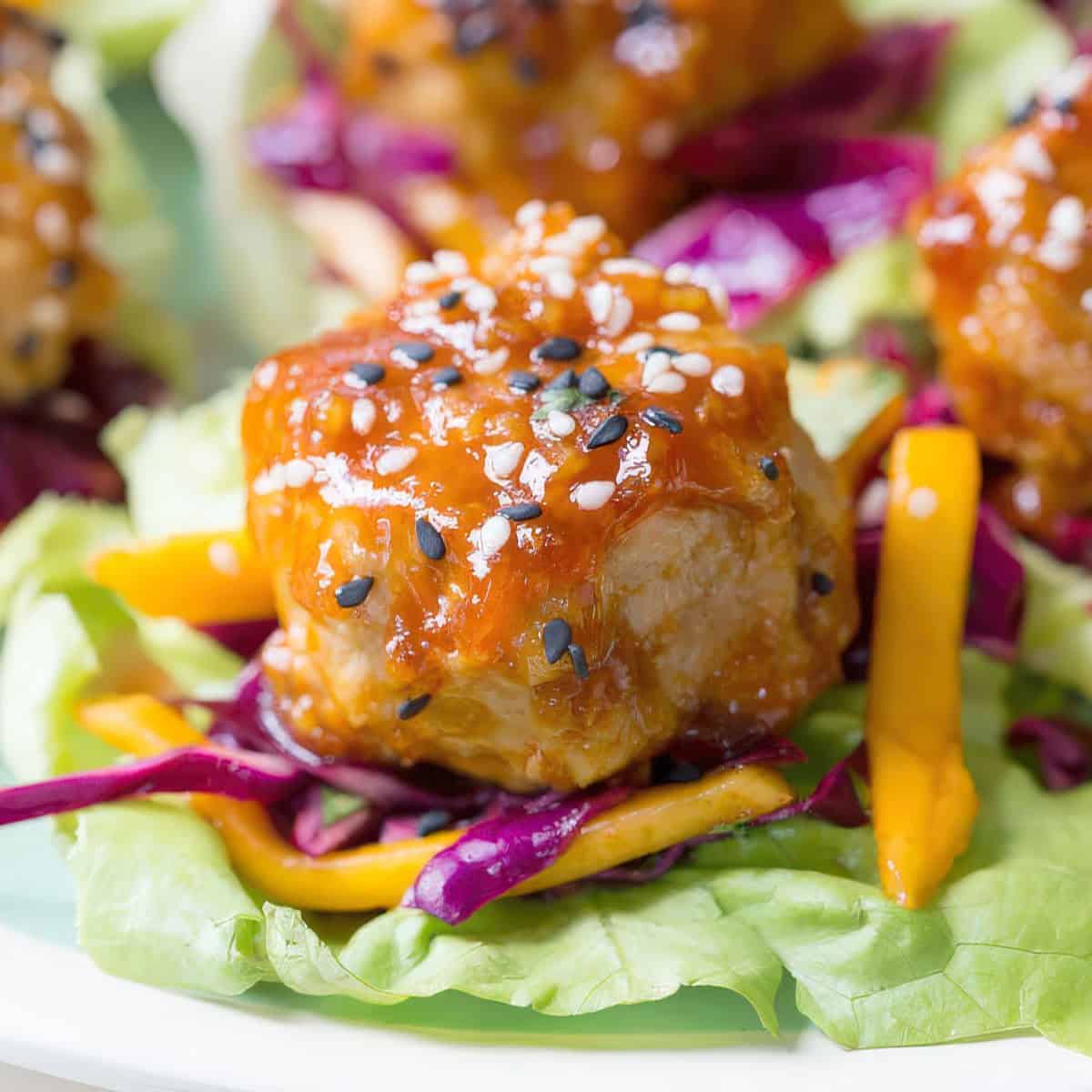 Close-up of a single lettuce wrap featuring a glazed chicken meatball, shredded red cabbage, mango slices, and a sprinkle of black and white sesame seeds on a butter lettuce leaf.