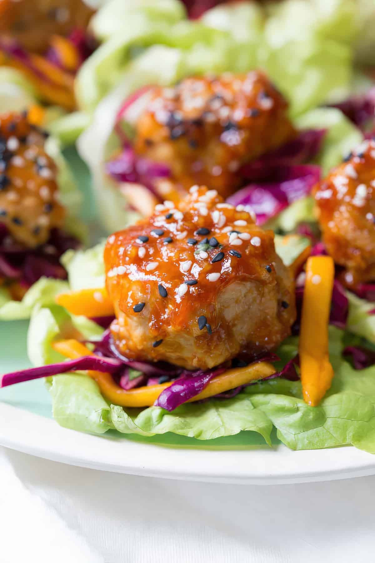 Close-up of a single lettuce wrap featuring a glazed chicken meatball, shredded red cabbage, mango slices, and a sprinkle of black and white sesame seeds on a butter lettuce leaf.