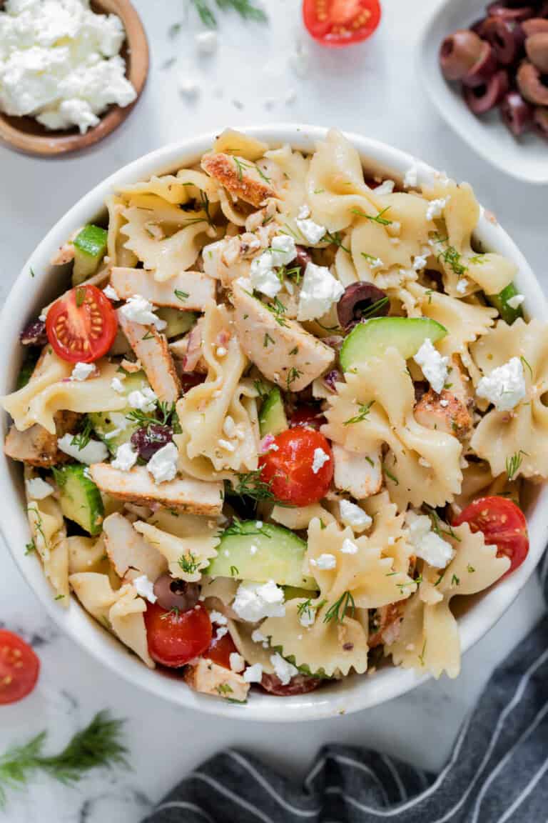Greek chicken pasta salad in a white bowl for serving. Cucumbers, tomatoes, kalamata olives, cooked chicken, bowtie pasta and feta cheese are piled high in the bowl.