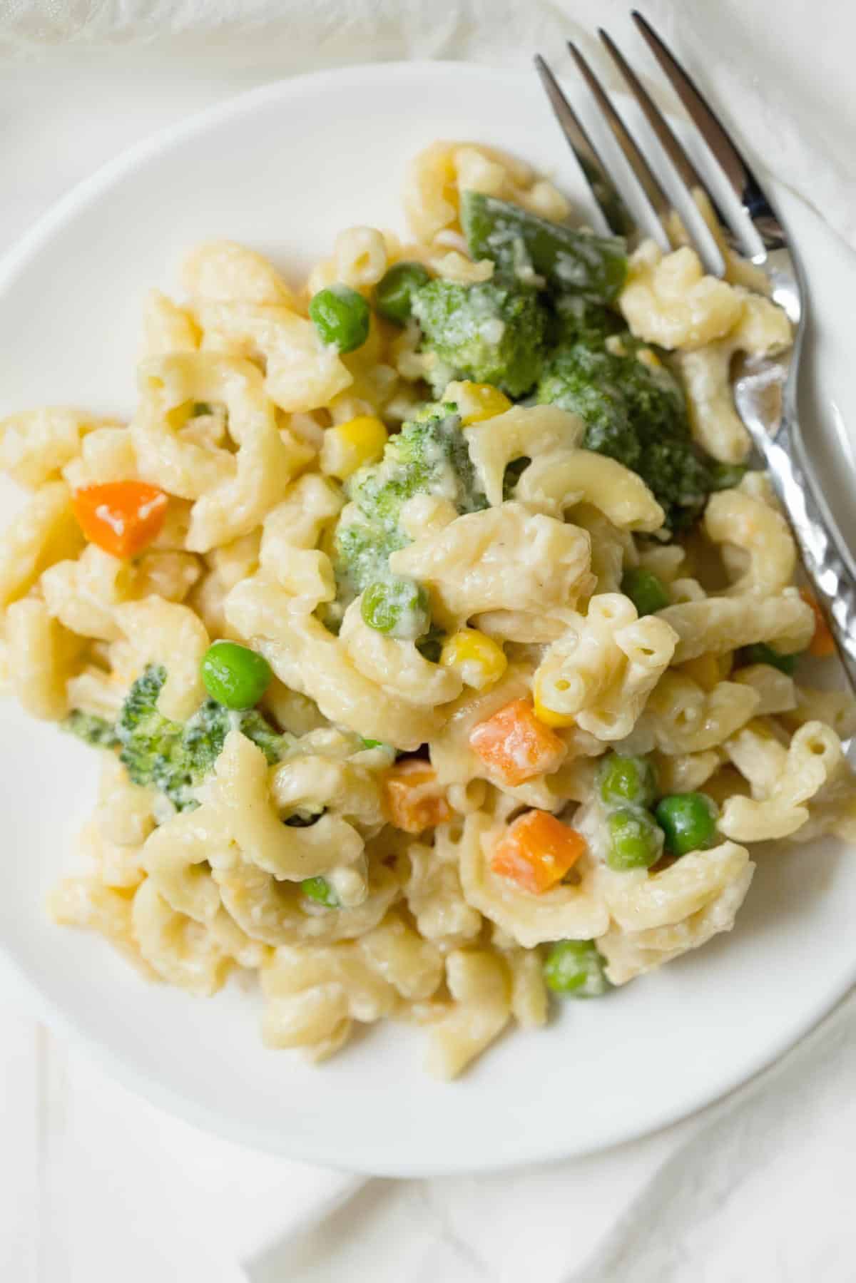 A serving of vegetable mac and cheese on a white plate with a silver fork, featuring colorful veggies and a creamy sauce.