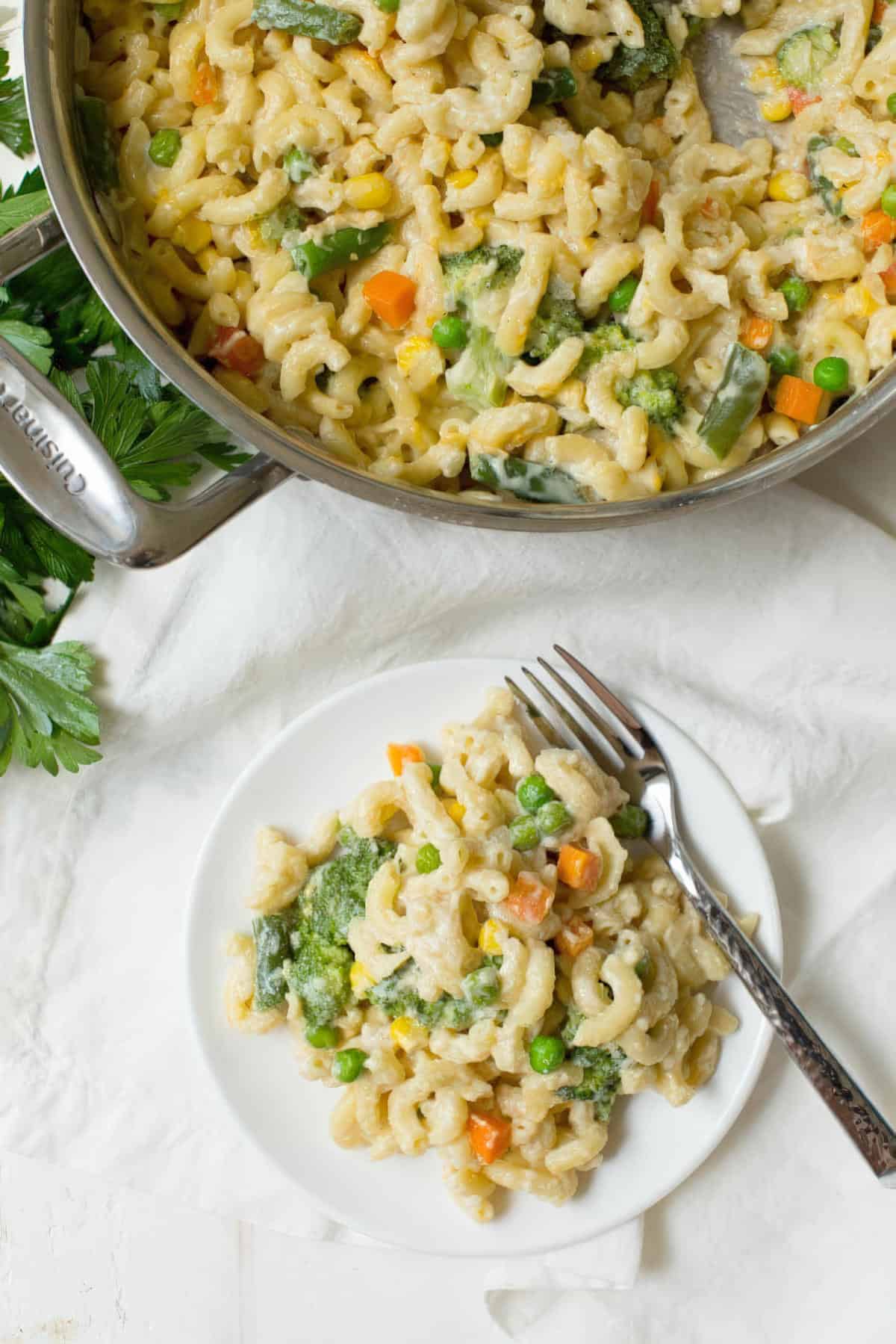 Cooked veggie mac and cheese in a pan with a serving spoon and a white plate of pasta next to it.