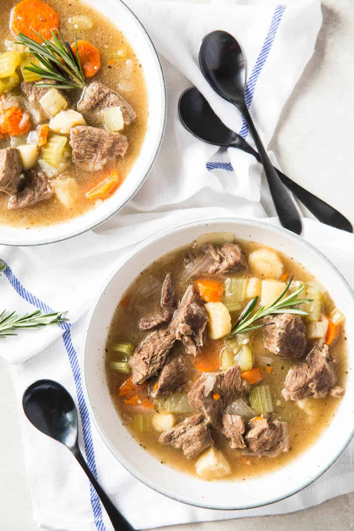 Two bowls of Instant Pot Beef Stew sit on a white tabletop with a napkin on the side and three spoons.