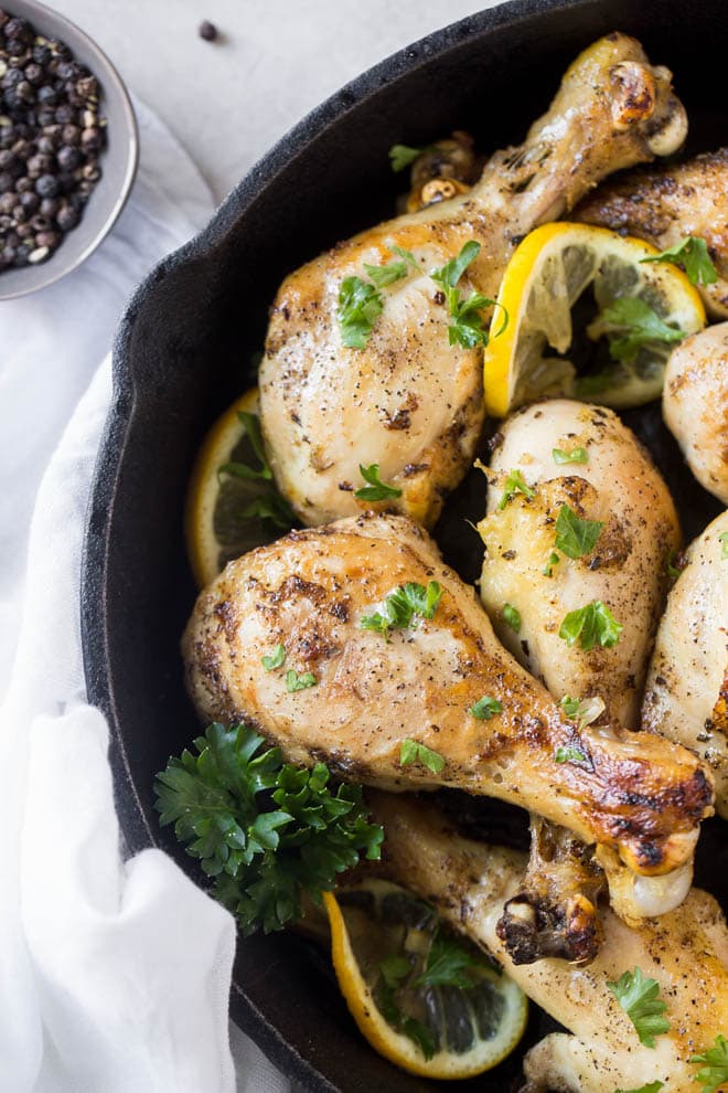 a few baked chicken legs in a black cast iron skillet with parsley garnish and lemon slices