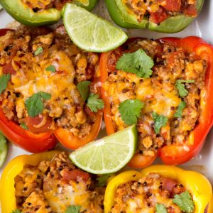 Six stuffed peppers sitting in a White dish with slices of lime and fresh parsley on top.