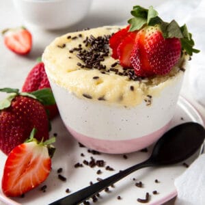 Vanilla mug cake with extra sprinkles and a sliced strawberry on top. A spoon sits on a plate on the side of the mug cake.