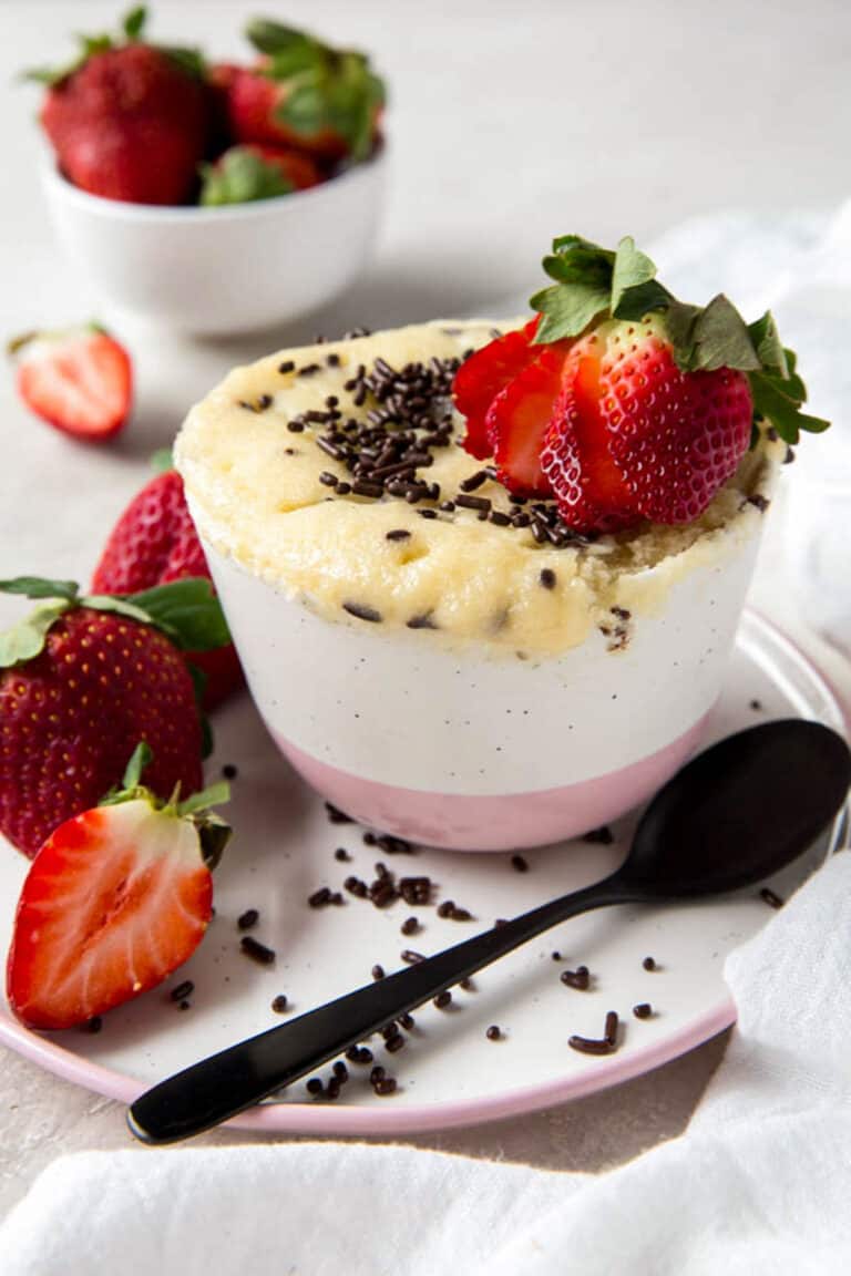 Vanilla mug cake with extra sprinkles and a sliced strawberry on top. A spoon sits on a plate on the side of the mug cake.