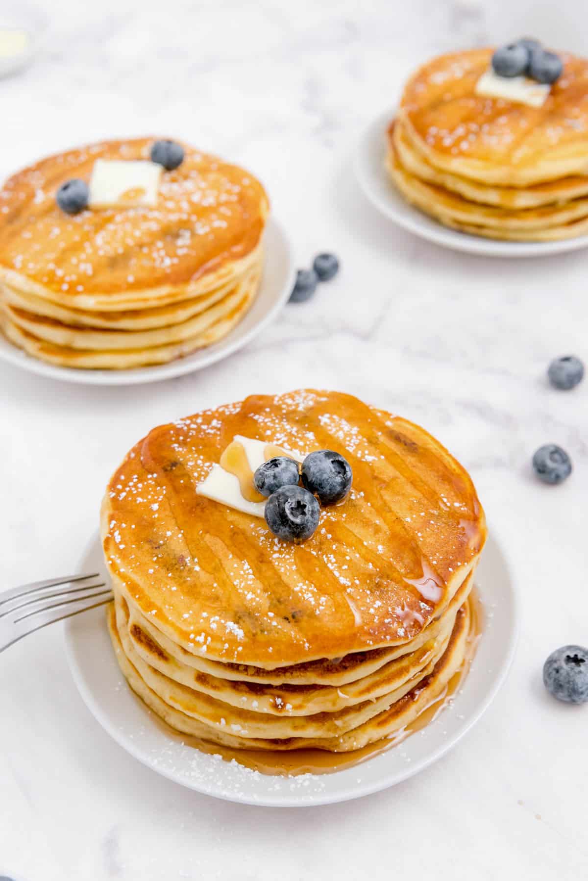 three stacks of blueberry buttermilk pancakes sitting on white plates on a counter
