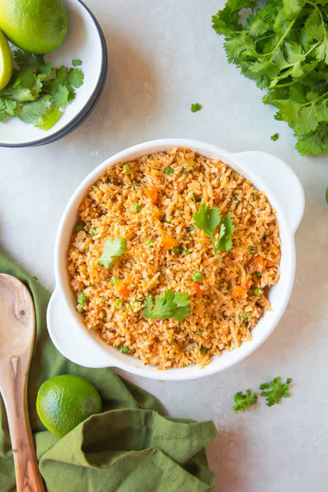 A large bowl of Mexican cauliflower rice sitting on a white table with a green napkin and wooden spoon off to the side. 