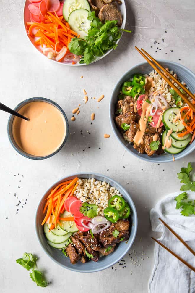 Two banh mi rice bowls sitting on a white backdrop with a bowl of sriracha mayo and a bowl of pickled veggies.
