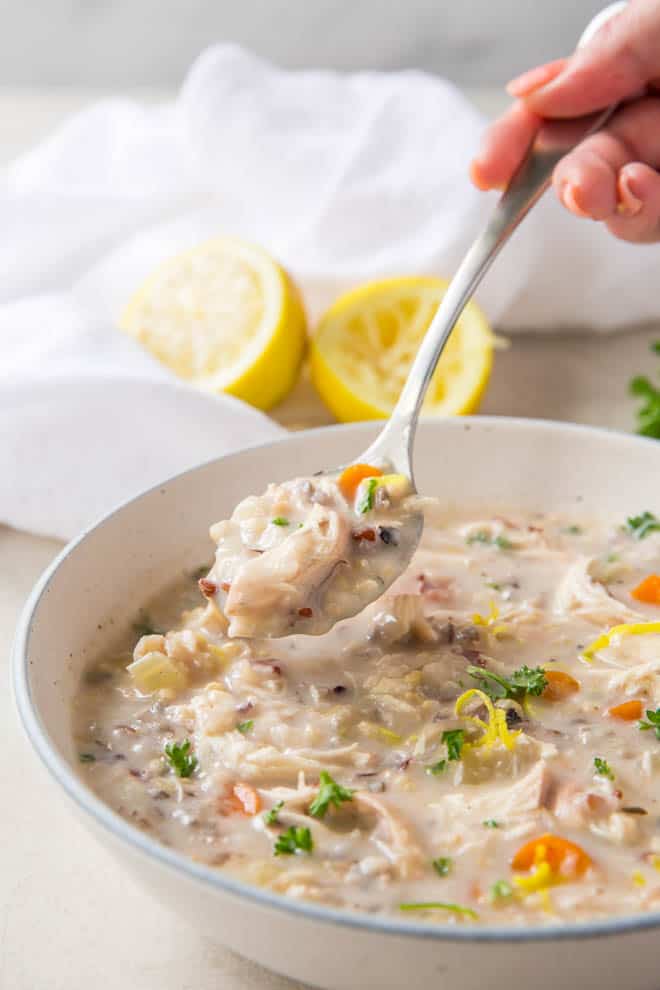 A spoon sits above a bowl of chicken and wild rice soup with two lemons in the background.