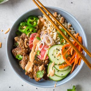 A bowl filled with brown rice, pork tenderloin, pickled veggies and sriracha mayo.