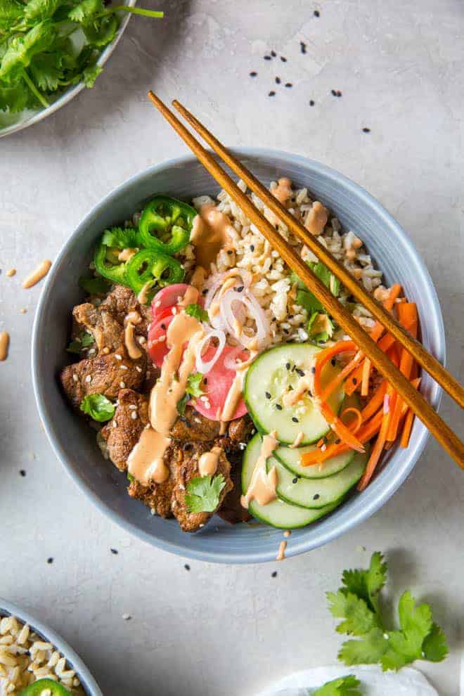 A bowl filled with brown rice, pork tenderloin, pickled veggies and sriracha mayo.