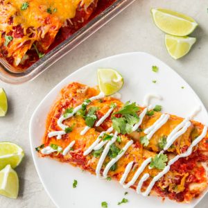 Two turkey enchiladas sit on a white plate with a casserole dish full of enchiladas off to the side.