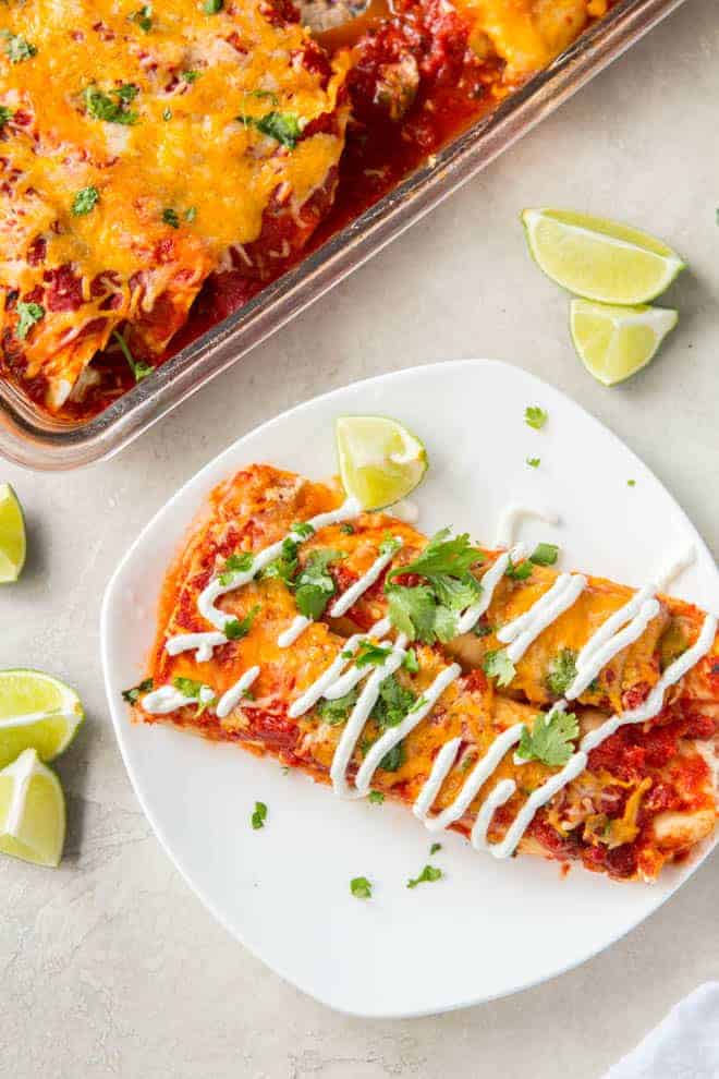 Two turkey enchiladas sit on a white plate with a casserole dish full of enchiladas off to the side. 